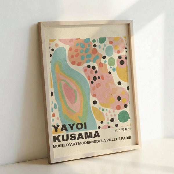 May include: Framed abstract art print with organic flowing shapes in soft pastels - teal, coral pink, yellow, and green circles and curved forms scattered across cream background. Black text reads "YAYOI KUSAMA" and "MUSEE D'ART MODERNE DE LA VILLE DE PARIS" with Japanese characters. Natural wood frame leaning against white wall.