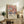 May include: Framed art print featuring colorful floral pattern in soft pastels - pink, coral, sage green, and navy flowers with radiating petal designs. Japanese text below artwork reads Yayoi Kusama with museum attribution. Print displayed on rustic wooden console table alongside fern plant, ceramic vase, and books in warm-lit living room with cream chair and neutral walls.