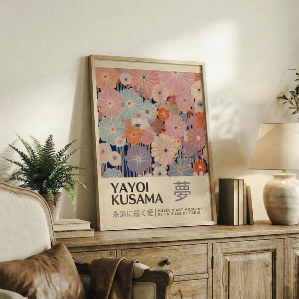 May include: Framed art print featuring colorful floral pattern in soft pastels - pink, coral, sage green, and navy flowers with radiating petal designs. Japanese text below artwork reads Yayoi Kusama with museum attribution. Print displayed on rustic wooden console table alongside fern plant, ceramic vase, and books in warm-lit living room with cream chair and neutral walls.