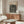 May include: Framed Yayoi Kusama exhibition poster with black polka dot pattern and wavy lines on cream background, displayed above mid-century modern walnut credenza in minimalist living room with white curved chair, glass coffee table, and brass floor lamp