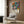May include: Colorful Yayoi Kusama exhibition poster with vibrant floral and polka dot patterns in blues, oranges, and pinks, displayed above a cream upholstered armchair with geometric pillow. Modern room setting features wood paneled walls, brass floor lamp, and dark round side table with open book.
