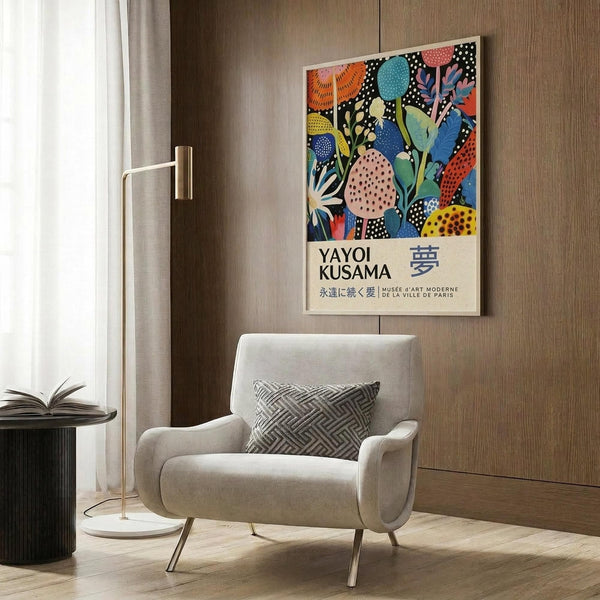 May include: Colorful Yayoi Kusama exhibition poster with vibrant floral and polka dot patterns in blues, oranges, and pinks, displayed above a cream upholstered armchair with geometric pillow. Modern room setting features wood paneled walls, brass floor lamp, and dark round side table with open book.
