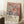 May include: A framed art print leaning against white walls on a textured cream rug, featuring colorful daisy-like flowers in pink, orange, mint green, and purple tones with dark blue stems against a navy background. The poster has cream-colored text area at bottom with "YAYOI KUSAMA" and Japanese characters, plus museum attribution text. A white coffee cup on wooden block and beige knit throw are visible nearby.