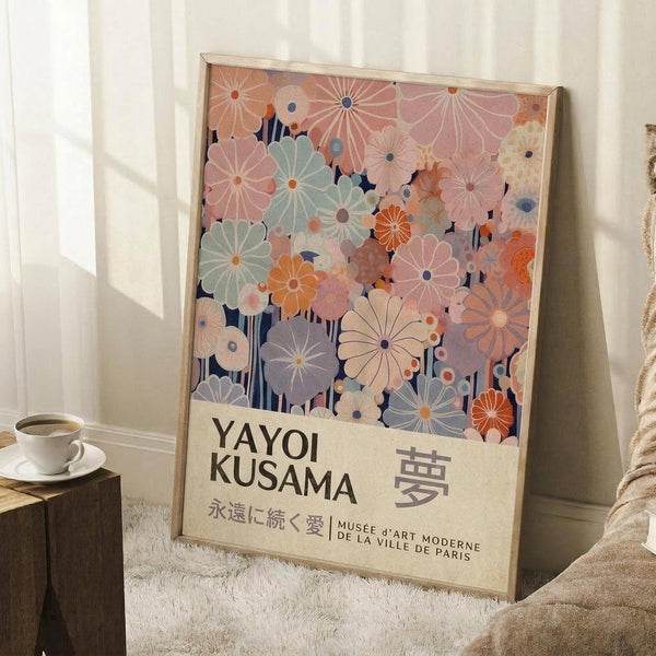 May include: A framed art print leaning against white walls on a textured cream rug, featuring colorful daisy-like flowers in pink, orange, mint green, and purple tones with dark blue stems against a navy background. The poster has cream-colored text area at bottom with "YAYOI KUSAMA" and Japanese characters, plus museum attribution text. A white coffee cup on wooden block and beige knit throw are visible nearby.