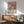 May include: Framed poster with colorful floral pattern in pink, orange, and sage green tones featuring Japanese text and museum attribution, displayed above mid-century modern wooden sideboard with white ceramic lamp, next to sage green upholstered armchair with wooden arms in sunlit room with white walls and wooden flooring.