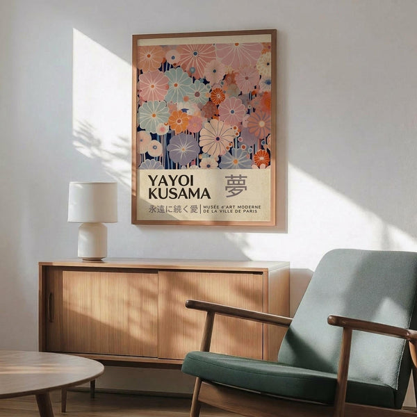May include: Framed poster with colorful floral pattern in pink, orange, and sage green tones featuring Japanese text and museum attribution, displayed above mid-century modern wooden sideboard with white ceramic lamp, next to sage green upholstered armchair with wooden arms in sunlit room with white walls and wooden flooring.