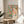 May include: Framed art poster with colorful abstract design featuring flowing teal shapes, pink and coral dots, black circles, and yellow accents on cream background. Text reads "YAYOI KUSAMA" and "MUSEE D'ART MODERNE DE LA VILLE DE PARIS" at bottom. Natural wood frame sits on round wooden side table next to beige upholstered chair. Soft natural lighting creates shadows on white wall. Green plant leaves visible in corner of minimalist room setting.