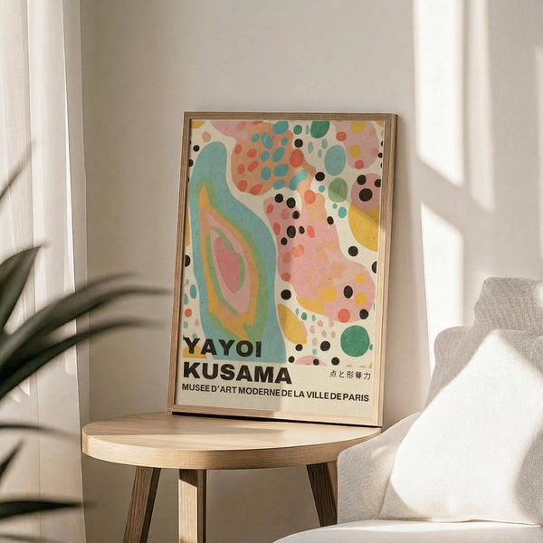 May include: Framed art poster with colorful abstract design featuring flowing teal shapes, pink and coral dots, black circles, and yellow accents on cream background. Text reads "YAYOI KUSAMA" and "MUSEE D'ART MODERNE DE LA VILLE DE PARIS" at bottom. Natural wood frame sits on round wooden side table next to beige upholstered chair. Soft natural lighting creates shadows on white wall. Green plant leaves visible in corner of minimalist room setting.
