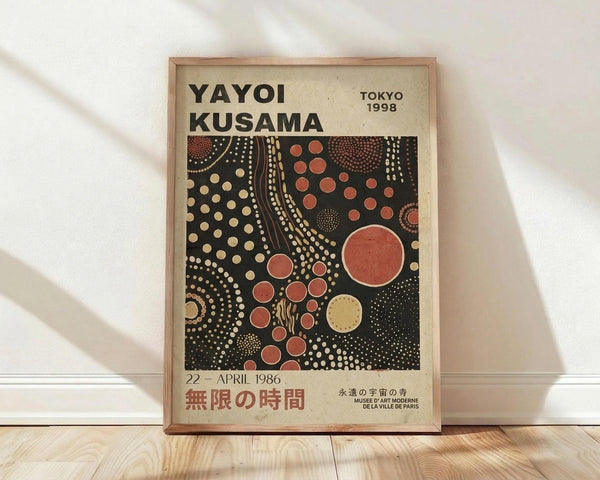 May include: Framed exhibition poster featuring Yayoi Kusama's signature polka dot artwork in black, coral red, and cream colors. The vintage-style poster displays "YAYOI KUSAMA" at the top with "TOKYO 1998" and Japanese text at the bottom. The central artwork shows flowing curved lines and various sized dots creating an abstract composition. The poster is displayed in a thin wooden frame against a white wall above light wooden flooring, with natural lighting creating soft shadows.