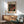May include: Framed Yayoi Kusama exhibition poster with black background featuring red, orange and cream polka dots in organic flowing patterns, displayed above a wooden sideboard with white ceramic lamp in a mid-century modern living room with mint green upholstered armchair and round wooden coffee table, natural sunlight casting shadows on white wall