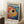 May include: Colorful abstract fish artwork in light wood frame leaning against white wall corner. Vibrant polka dot style painting features large fish with prominent black eye surrounded by rainbow circles and dots in red, blue, yellow, orange hues. Japanese text at top and bottom. Small wooden side table with white coffee cup visible beside framed print on textured cream rug.