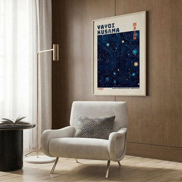 May include: Framed poster with deep blue cosmic design featuring swirling patterns and colorful dots resembling planets, displayed on wood-paneled wall above modern beige armchair with geometric pillow. Golden floor lamp and dark round side table with open book visible in warm, contemporary living room setting with natural light from window.