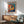 May include: Framed colorful abstract fish artwork with dotted pattern and large eye, displayed above wooden credenza with white lamp in modern living room setting. Green upholstered armchair with wooden frame sits nearby. Natural sunlight creates shadows on white wall. Art features vibrant rainbow colors transitioning from warm oranges and reds to cool blues, covered in polka dots in Kusama's signature infinity style.