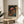 May include: Framed art print featuring colorful mushrooms in various sizes against a dark starry background, displayed on a round wooden side table next to a beige upholstered chair. The artwork shows vibrant red, orange, yellow and blue fungi with polka dot caps and long stems, surrounded by floating dots and circles. Natural lighting creates soft shadows on white walls, with a plant leaf visible in the corner.