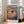 May include: Framed colorful abstract fish artwork in light wood frame leaning against white wall next to beige sofa. Vibrant poster features stylized fish with large dark eye surrounded by rainbow dots and swirling patterns in red, orange, yellow, blue, and green. Black text at top, Japanese characters at bottom. Bright natural lighting from tall windows creates soft shadows on wooden floor.