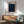 May include: Framed poster with deep blue starry night sky design featuring swirling patterns and scattered celestial orbs in blues and yellows, displayed above wooden sideboard with ceramic table lamp. Mid-century modern sage green armchair with wooden arms sits nearby on hardwood floor with natural sunlight casting shadows across white wall.