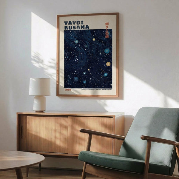 May include: Framed poster with deep blue starry night sky design featuring swirling patterns and scattered celestial orbs in blues and yellows, displayed above wooden sideboard with ceramic table lamp. Mid-century modern sage green armchair with wooden arms sits nearby on hardwood floor with natural sunlight casting shadows across white wall.