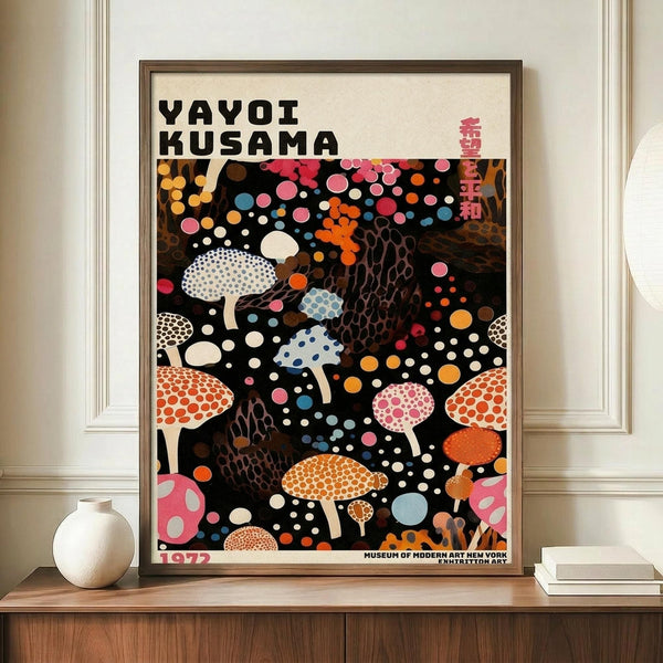 May include: Framed poster featuring colorful polka-dotted mushrooms on black background with scattered circles in pink, orange, blue, and white. Text reads "YAYOI KUSAMA" and "1972" with Japanese characters. Brown wooden frame displayed on light wood surface beside white ceramic vase and stacked books against cream wall with white trim molding.