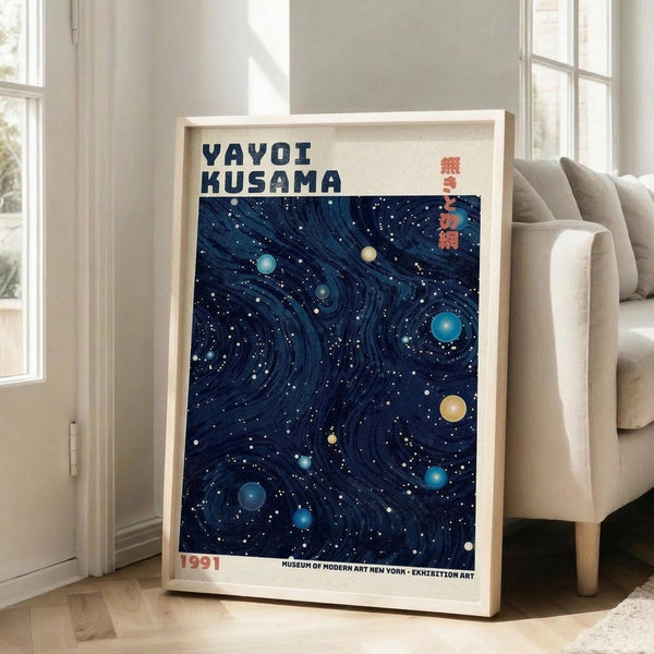 May include: Framed art print with deep blue swirling background filled with white dots and colorful circular planets in yellow, turquoise, and blue tones. Bold black text reads "YAYOI KUSAMA" at top with red Japanese characters and "1991" at bottom. Light wood frame leans against white wall next to beige upholstered chair in bright, minimalist living room with large windows.