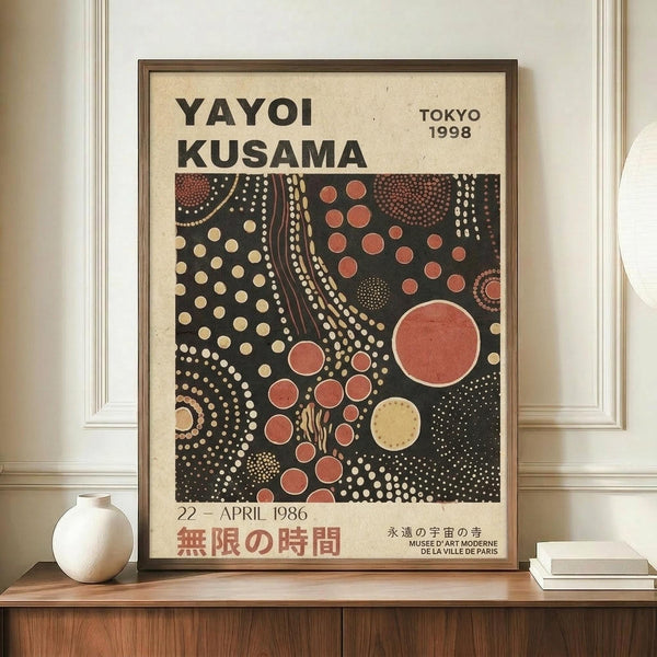 May include: Framed exhibition poster featuring Yayoi Kusama's signature polka dot artwork in black background with coral red, cream and yellow circular patterns and flowing dotted lines. Text shows Tokyo 1998 and April 1986 dates with Japanese characters. Displayed on wooden console table beside white ceramic vase and books against cream walls with white trim molding.