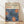 May include: Framed art print featuring stylized blue flowers with orange dotted centers on cream background, displayed in light wood frame against white wall on wooden floor with natural window light creating soft shadows