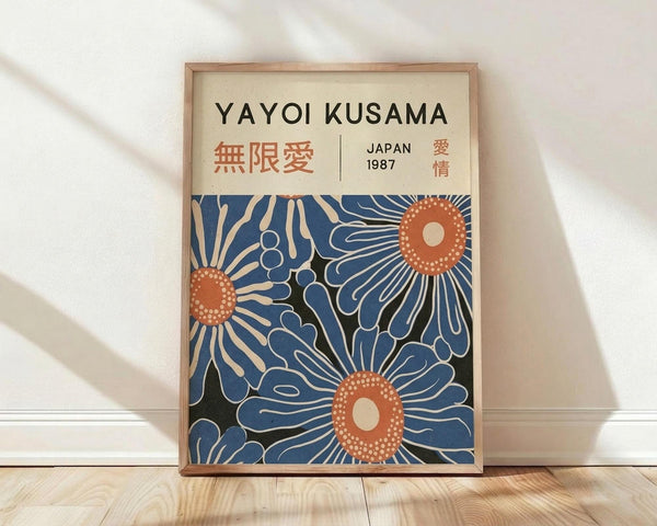 May include: Framed art print featuring stylized blue flowers with orange dotted centers on cream background, displayed in light wood frame against white wall on wooden floor with natural window light creating soft shadows