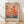May include: Framed poster print leaning against white wall on wooden floor. Artwork features abstract forest of colorful circular tree-like forms in warm reds, oranges, and yellows transitioning to cool blues at bottom. Polka dot patterns fill many of the round shapes on thin stems. Text in Japanese characters and "YAYOI KUSAMA" appears at bottom in red letters on cream background. Natural wood frame, soft lighting creates diagonal shadows on white textured wall.