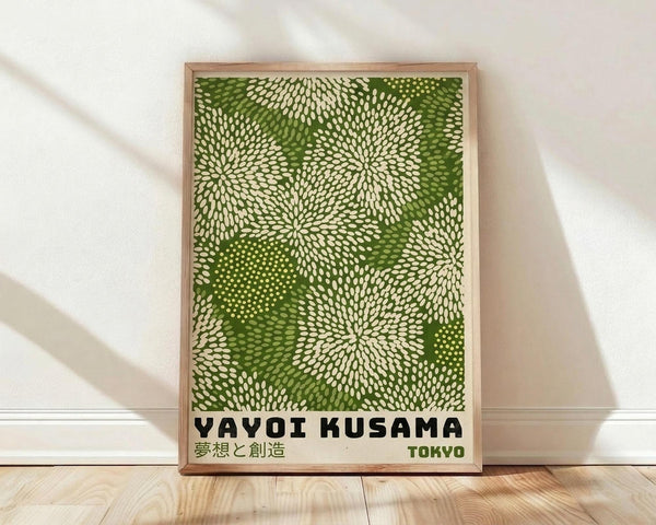 May include: Framed poster with green and cream dot pattern forming radiating flower-like designs, displayed against white wall on wooden floor. Natural wood frame surrounds artwork featuring "YAYOI KUSAMA" and "TOKYO" text at bottom with Japanese characters. Soft natural lighting creates diagonal shadows across the scene.