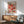 May include: Framed Yayoi Kusama exhibition poster from 1981 featuring vibrant polka-dotted organic shapes in coral, pink, orange, and teal tones, displayed above a mid-century modern wooden sideboard with white ceramic lamp, next to a sage green upholstered armchair with wooden frame in a sunlit room with white walls
