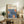 May include: Framed art print with cream background featuring blue floral design and orange circular elements, displayed on rustic wooden sideboard alongside decorative pottery, books, and fern plant in neutral-toned living room setting with beige upholstered furniture