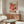 May include: Colorful Yayoi Kusama exhibition poster with vibrant polka dot pattern in pink, red, orange, and green tones, displayed above mid-century modern wooden credenza with sliding doors. White sculptural chair and glass-top oval coffee table visible in minimalist living room with cream walls and natural lighting.