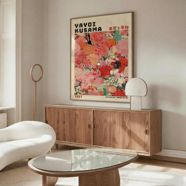 May include: Colorful Yayoi Kusama exhibition poster with vibrant polka dot pattern in pink, red, orange, and green tones, displayed above mid-century modern wooden credenza with sliding doors. White sculptural chair and glass-top oval coffee table visible in minimalist living room with cream walls and natural lighting.