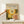 May include: Hand holding wooden-framed poster featuring colorful circular dot patterns in yellow, orange, and green tones on dark background, displayed on light wood furniture next to small white wrapped gift box against cream wall