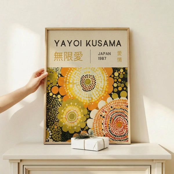 May include: Hand holding wooden-framed poster featuring colorful circular dot patterns in yellow, orange, and green tones on dark background, displayed on light wood furniture next to small white wrapped gift box against cream wall