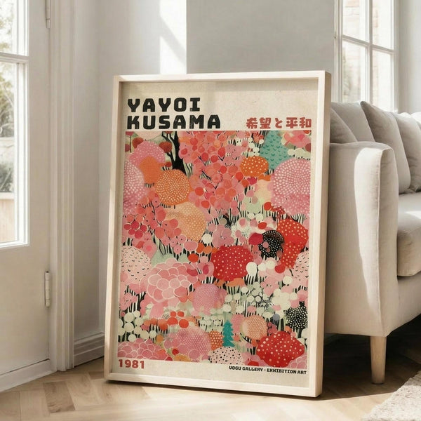 May include: Framed art print leaning against white wall next to beige upholstered chair in bright living room. Poster features colorful abstract pattern of overlapping circular shapes in coral, pink, orange, mint green, and black with polka dots and geometric textures. Black text at top with Japanese characters, date 1981 at bottom. Light wood frame, cream matting. Natural lighting from windows creates soft shadows on white walls and light wooden floors.