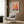 May include: Colorful abstract art poster with polka dot balloon-like circles in red, orange, pink, and blue tones, displayed in white frame on wood-paneled wall above beige upholstered chair with geometric pillow. Modern reading corner features brass floor lamp and dark round side table with open book. Warm neutral interior with natural lighting from window.