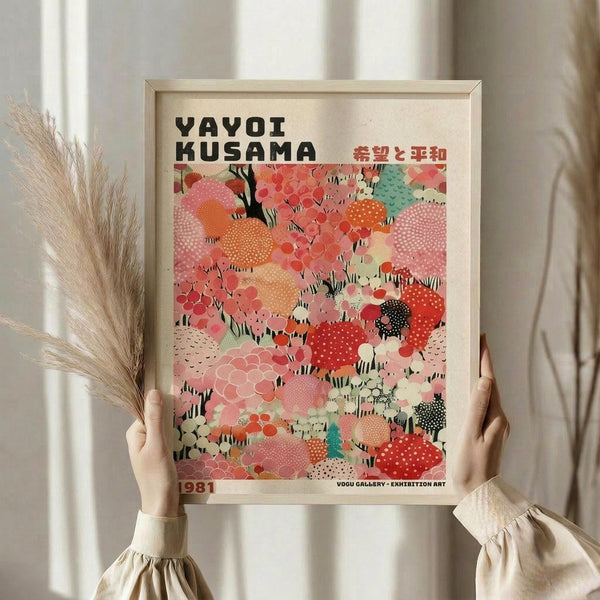 May include: Person in beige ruffled sleeves holding a framed art poster featuring colorful abstract polka dot patterns in coral, pink, orange, and mint green with black tree branches. The poster displays "YAYOI KUSAMA" at the top with Japanese characters and "1981" at the bottom. Light wood frame, neutral background with dried pampas grass visible to the left.