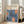 May include: Framed art print leaning against white wall next to beige upholstered chair in bright living room. Poster features blue and orange floral design with three large daisy-like flowers, white petals, orange centers with dotted patterns. Text reads "YAYOI KUSAMA" at top with Japanese characters and "JAPAN 1987". Light wood frame, cream background, natural sunlight streaming through windows onto hardwood floors.