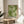 May include: Framed green and cream poster with dotted floral pattern displayed on light wood round side table next to beige upholstered chair. Natural sunlight streams across white walls creating soft shadows. Green plant leaves visible in corner. Poster features Japanese text and "TOKYO" at bottom in thin wooden frame.