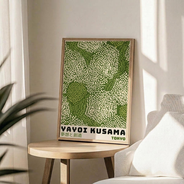 May include: Framed green and cream poster with dotted floral pattern displayed on light wood round side table next to beige upholstered chair. Natural sunlight streams across white walls creating soft shadows. Green plant leaves visible in corner. Poster features Japanese text and "TOKYO" at bottom in thin wooden frame.