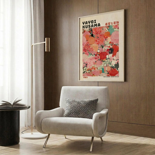 May include: Colorful Yayoi Kusama exhibition poster featuring abstract polka dot patterns in coral, pink, orange, and teal tones, displayed in light wood frame on wood paneled wall above modern beige armchair with geometric pillow, brass floor lamp, and side table with open book in contemporary room setting.