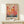 May include: Framed art print featuring abstract colorful circles and dots in warm reds, oranges, and yellows with cool blues at bottom, resembling floating balloons or organic forms with Japanese text below. Light wood frame displayed on white wooden dresser beside small white wrapped gift with evergreen sprig against cream wall.