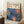May include: Large framed art print leaning against white wall corner on textured white rug, featuring blue floral design with orange dotted centers and Japanese text at top. Natural wood frame, nearby coffee cup on wooden block, soft natural lighting creates shadows on wall.