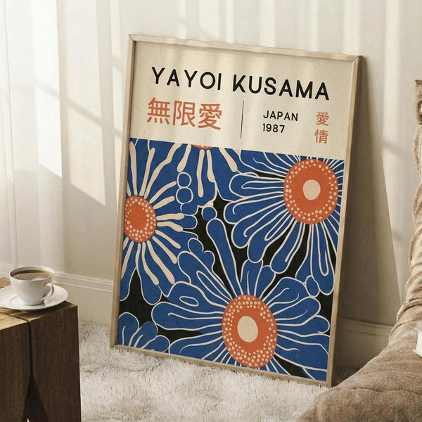 May include: Large framed art print leaning against white wall corner on textured white rug, featuring blue floral design with orange dotted centers and Japanese text at top. Natural wood frame, nearby coffee cup on wooden block, soft natural lighting creates shadows on wall.
