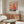 May include: Colorful abstract art poster with polka dot patterns in red, orange, blue and yellow tones, displayed in thin gold frame above wooden credenza. Modern living room setting features white curved chair, glass oval coffee table, and contemporary table lamp. Warm natural lighting creates shadows on light gray walls.