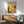May include: Framed art poster with colorful circular dot patterns in yellow, orange, and green tones displayed on white wall above wooden sideboard with lamp, beside mid-century modern teal upholstered chair with wooden arms in bright, naturally lit room with contemporary furniture
