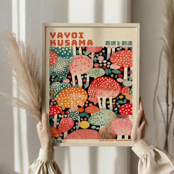 May include: Person holding framed colorful mushroom art print in natural wood frame. Vibrant illustration features polka-dotted mushrooms in red, orange, pink, teal, and yellow against black background. Japanese text at top reads "YAYOI KUSAMA" with green characters below. Dried pampas grass visible to left, neutral beige clothing and curtains in soft natural lighting.