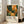 May include: Framed art print leaning against white wall next to beige sofa in bright living room. Poster features vibrant illustration with orange background, green foliage, yellow mushrooms, and white polka dots in Kusama's distinctive style. White frame surrounds artwork with text reading "YAYOI KUSAMA" and Japanese characters below. Natural lighting from windows creates soft shadows on wooden floor.
