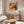 May include: Framed colorful abstract floral poster featuring large yellow dahlia, pink and orange circular patterns, and black polka dot elements, displayed above mid-century walnut credenza in minimalist living room with white curved chair, glass oval coffee table, and brass floor lamp