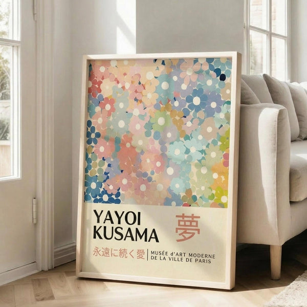 May include: Framed art print featuring colorful abstract polka dot pattern in pastel blues, pinks, corals and yellows with "YAYOI KUSAMA" text and Japanese characters below, leaning against white wall next to beige upholstered chair in bright sunlit room with large windows and wooden floors.